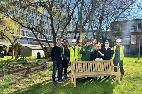 A group of people standing behind a wooden bench in a garden.