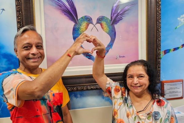 Rasik and Sarla making a heart shape with their hands, in front of a painting of two humming birds with their beaks an tails together in a heart shape.